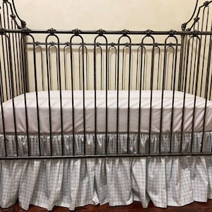 Pu&ograve; includere: Una culla in metallo scuro con un materasso bianco e una gonna per culla a quadretti vichy blu chiaro e bianco. La culla presenta decorazioni a volute sulla testiera e sulla pediera e barre verticali sui lati.