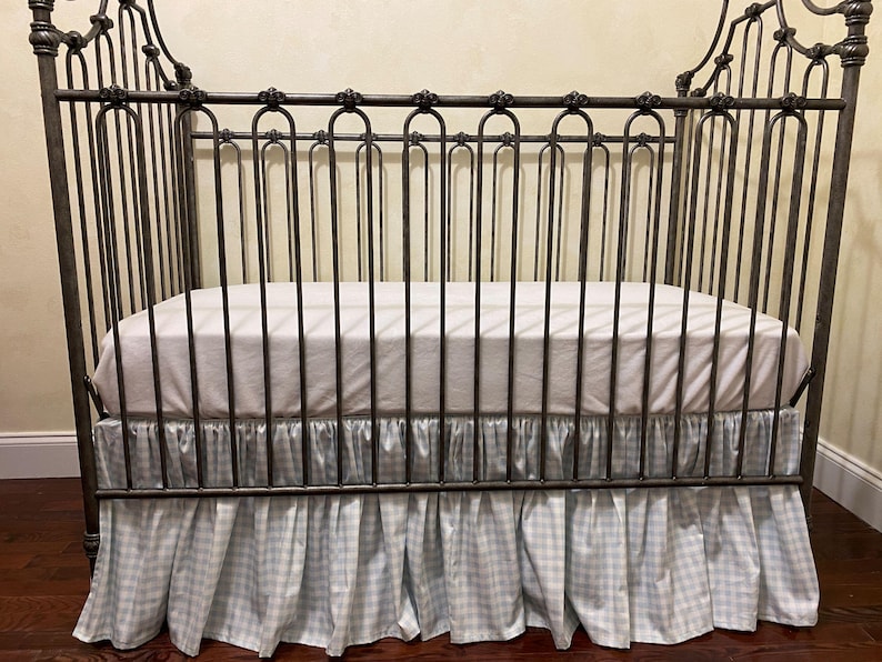Pu&ograve; includere: Una culla in metallo scuro con un materasso bianco e una gonna per culla a quadretti vichy blu chiaro e bianco. La culla presenta decorazioni a volute sulla testiera e sulla pediera e barre verticali sui lati.