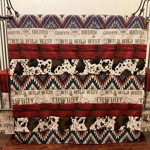 May include: A red, white, and blue crib bedding set with a cowboy theme. The bedding features a patchwork design with cowhide print, wooden planks, and geometric patterns. The text "Giddyup Rodeo Wild Wild West Cowboy" is printed on the bedding.
