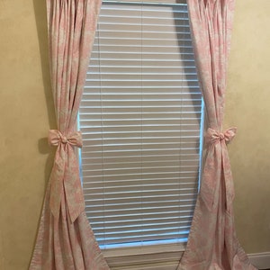 May include: A pair of pink curtains with a white floral pattern, hung on a window with white blinds. The curtains are tied back with bows.
