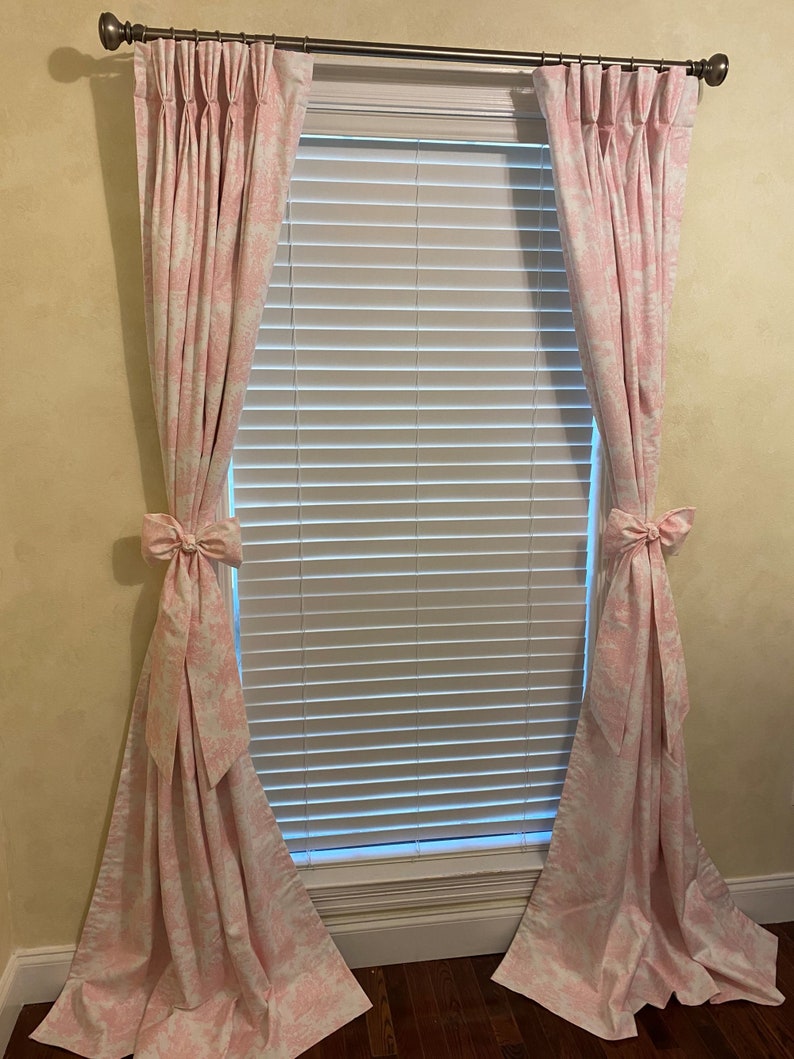 May include: A pair of pink curtains with a white floral pattern, hung on a window with white blinds. The curtains are tied back with bows.