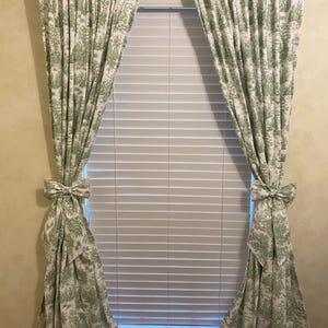 May include: A pair of green and white patterned curtains with a bow tie detail at the bottom. The curtains are hung on a metal rod and are covering a window with white blinds.
