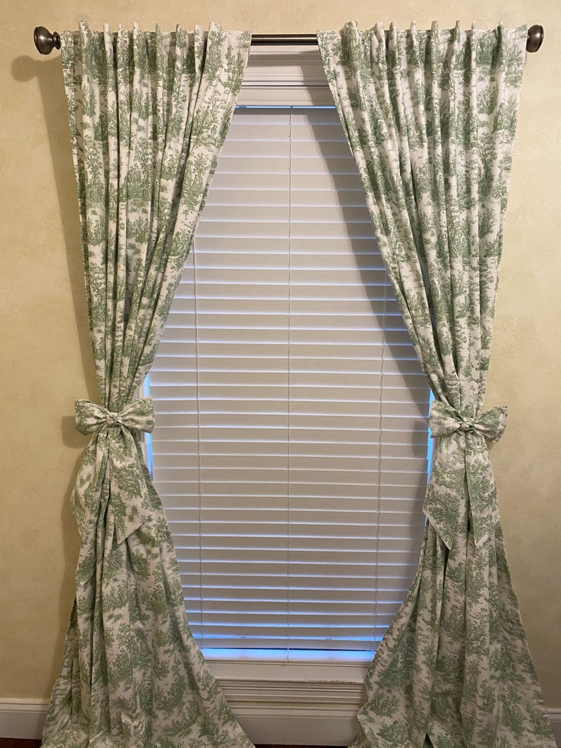 May include: A pair of green and white patterned curtains with a bow tie detail at the bottom. The curtains are hung on a metal rod and are covering a window with white blinds.