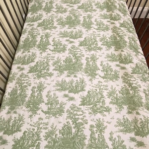 Op de afbeelding: Een wit babybeddengoed met een groen toile-patroon met een herhalend ontwerp van bomen, mensen en dieren.