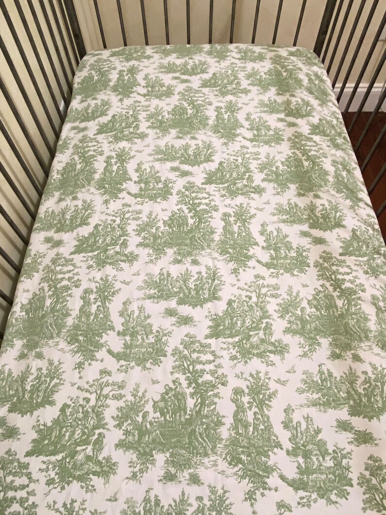 Op de afbeelding: Een wit babybeddengoed met een groen toile-patroon met een herhalend ontwerp van bomen, mensen en dieren.