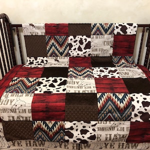 May include: A patchwork baby quilt with a cowboy theme. The quilt is made of red, brown, white, and blue fabrics with a variety of patterns, including cowhide, chevron, and wood grain. The quilt features the text "Boy Wild We Round Em Up" and "Ye Haw Cowboy".