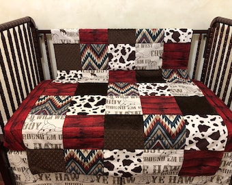 May include: A patchwork baby quilt with a cowboy theme. The quilt is made of red, brown, white, and blue fabrics with a variety of patterns, including cowhide, chevron, and wood grain. The quilt features the text "Boy Wild We Round Em Up" and "Ye Haw Cowboy".