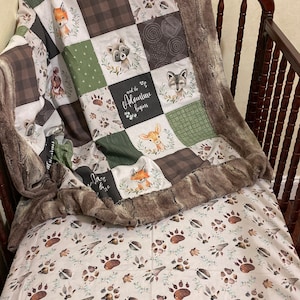 May include: A brown and green baby blanket with a patchwork design featuring woodland animals and the text "and the adventure begins". The blanket is on a crib with a white sheet with a brown paw print pattern.