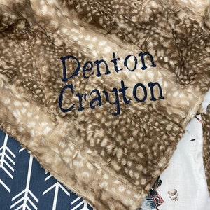 May include: A plush, brown and beige blanket with a spotted pattern, embroidered with the name "Denton Crayton" in navy blue thread. The blanket is folded over other patterned fabrics.
