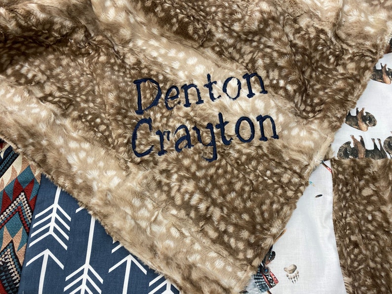 May include: A plush, brown and beige blanket with a spotted pattern, embroidered with the name "Denton Crayton" in navy blue thread. The blanket is folded over other patterned fabrics.