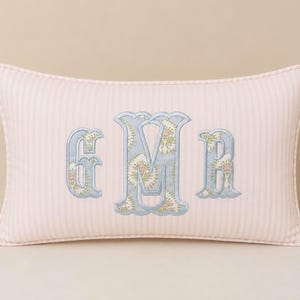 May include: A rectangular throw pillow featuring a light pink and white vertical striped fabric. The center displays a large embroidered monogram with the letters G, M, and R in a light blue floral pattern with a light blue stitched border.