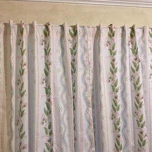 May include: White floral patterned curtains with green leaves, pink flowers, and blue wavy lines. The curtains are hanging on a dark brown rod, adding a touch of elegance to the room. The design is repeated vertically.