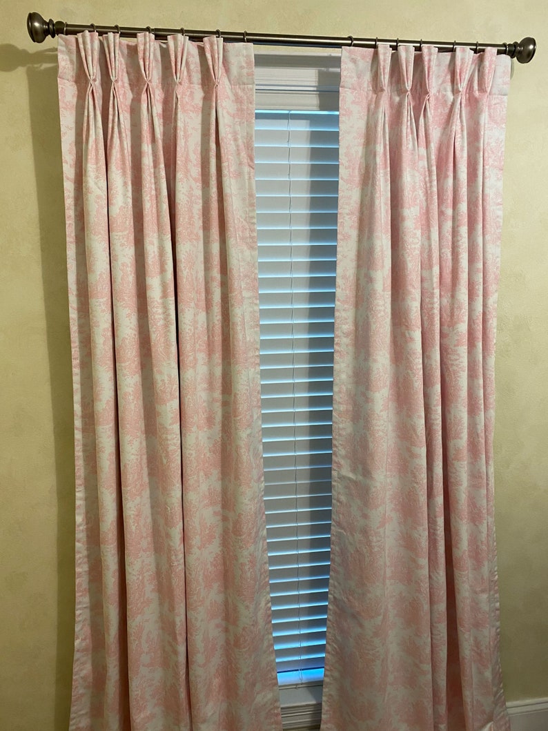 May include: A pair of pink and white patterned curtains with pleats hanging in a window with white blinds.