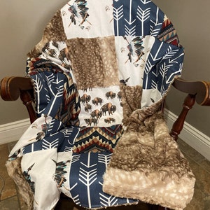 May include: A patchwork blanket draped over a wooden chair. The blanket features various patterns, including white fabric with Native American headdresses, navy blue arrows, and brown faux fur. The blanket's design includes bison and geometric patterns.