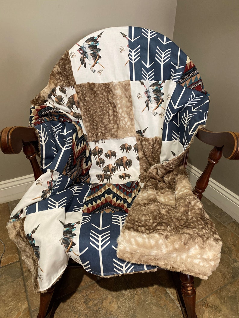 May include: A patchwork blanket draped over a wooden chair. The blanket features various patterns, including white fabric with Native American headdresses, navy blue arrows, and brown faux fur. The blanket's design includes bison and geometric patterns.