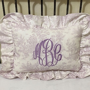 May include: A white pillow with a lavender floral pattern and a lavender monogram "RE" in the center. The pillow has a ruffled edge.