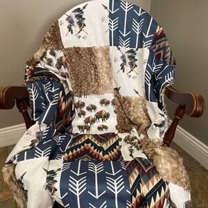 May include: A patchwork blanket draped over a wooden chair. The blanket features various patterns, including bison, Native American headdresses, arrows, and geometric designs in shades of brown, white, and navy blue. The blanket has a faux fur trim.