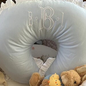 May include: A light blue nursing pillow with white embroidered initials and a ruffled edge. The pillow is in a basket with a teddy bear and a stuffed chick. A baby item.