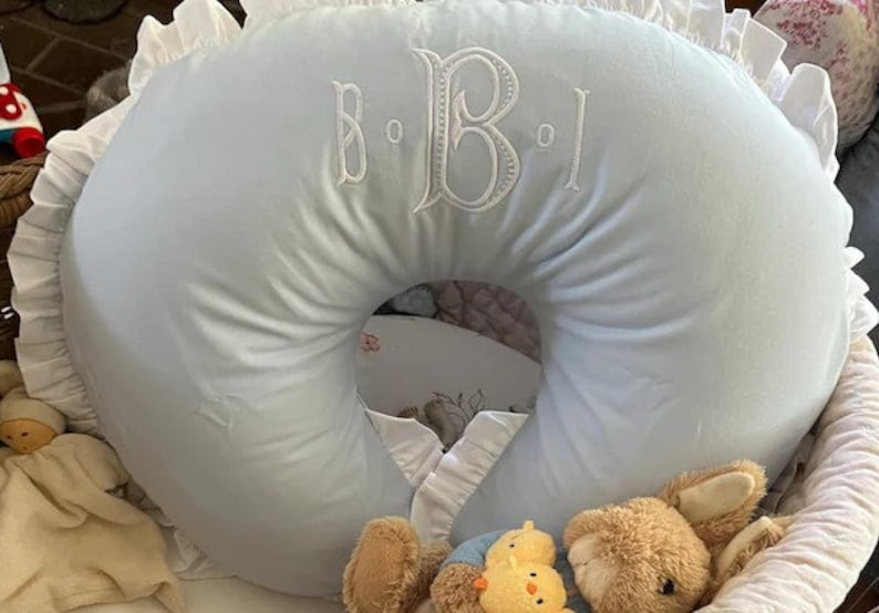 May include: A light blue nursing pillow with white embroidered initials and a ruffled edge. The pillow is in a basket with a teddy bear and a stuffed chick. A baby item.