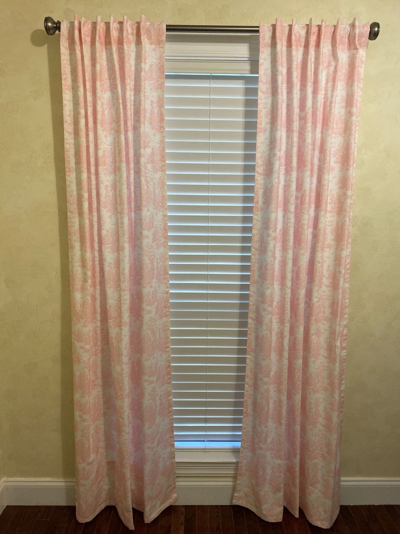 May include: A pair of pink and white patterned curtains hang in front of a window with white blinds. The curtains are made of a lightweight fabric and have a subtle sheen.