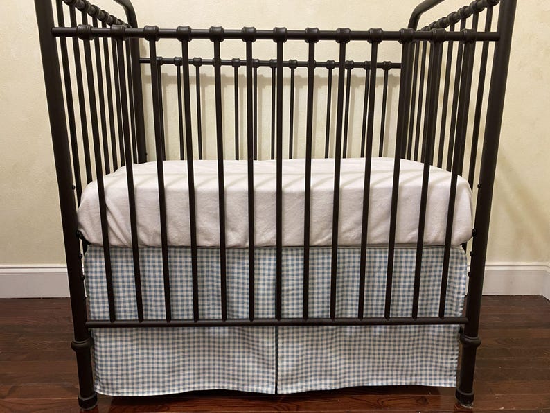 Puede incluir: Una cuna de metal marr&oacute;n oscuro con barras verticales y un colch&oacute;n blanco. La cuna tiene una falda con estampado de cuadros vichy azul claro y blanco. La cuna est&aacute; dise&ntilde;ada para un beb&eacute; o ni&ntilde;o peque&ntilde;o.