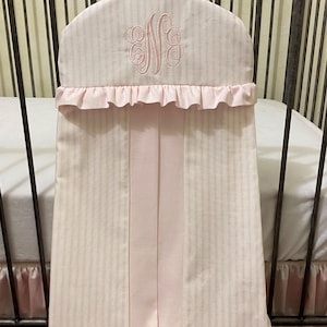 May include: A pink and white striped crib bumper with a ruffled edge and a monogram. The bumper is hanging on a metal crib frame. The crib skirt is also pink and white striped.