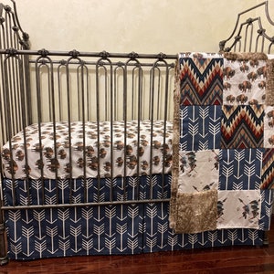 May include: A crib with a metal frame, featuring bedding with a Native American theme. The fitted sheet and crib skirt have arrow patterns, while the blanket showcases bison and geometric designs in shades of brown, blue, and red.