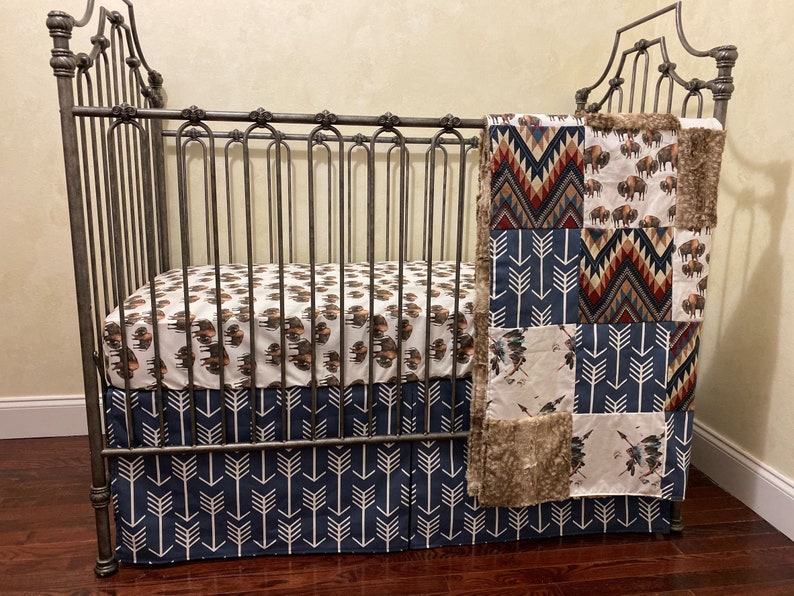 May include: A crib with a metal frame, featuring bedding with a Native American theme. The fitted sheet and crib skirt have arrow patterns, while the blanket showcases bison and geometric designs in shades of brown, blue, and red.