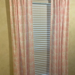 May include: A pair of pink and white patterned curtains hang in front of a window with white blinds. The curtains have a delicate floral design.