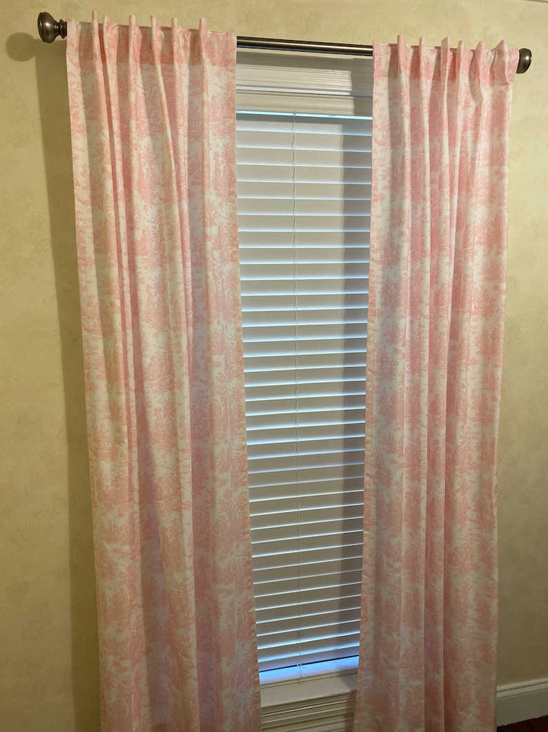May include: A pair of pink and white patterned curtains hang in front of a window with white blinds. The curtains have a delicate floral design.