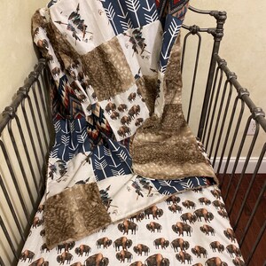 May include: A patchwork quilt with a Native American theme, featuring bison, arrows, and tribal patterns in shades of brown, navy, and cream. The quilt is draped over a dark metal crib frame, with a faux fur trim.
