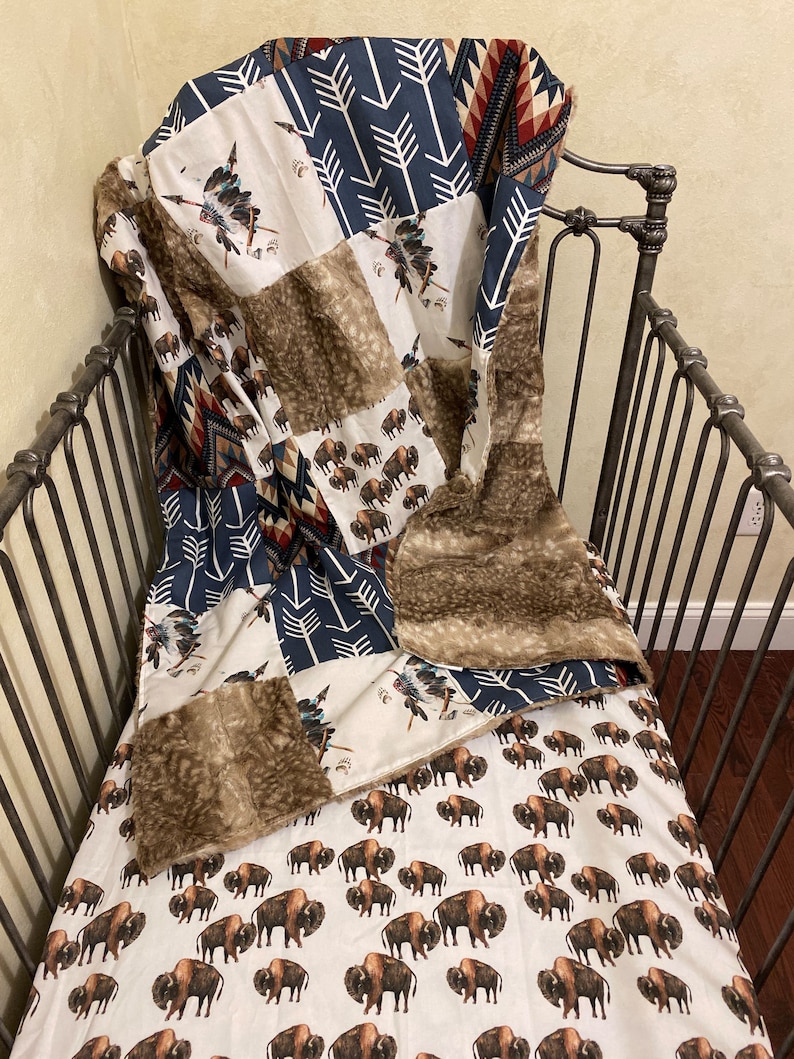 May include: A patchwork quilt with a Native American theme, featuring bison, arrows, and tribal patterns in shades of brown, navy, and cream. The quilt is draped over a dark metal crib frame, with a faux fur trim.