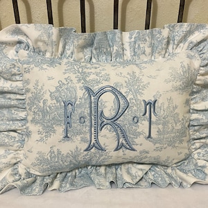 May include: A white pillow with a blue toile pattern and a blue embroidered monogram "F.R.T." on the front. The pillow has a ruffled edge.