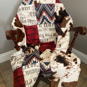 May include: A patchwork quilt with a cowboy theme. The quilt features a red, white, and blue color scheme with a variety of patterns, including cowhide, chevron, and woodgrain. The quilt is draped over a rocking chair.