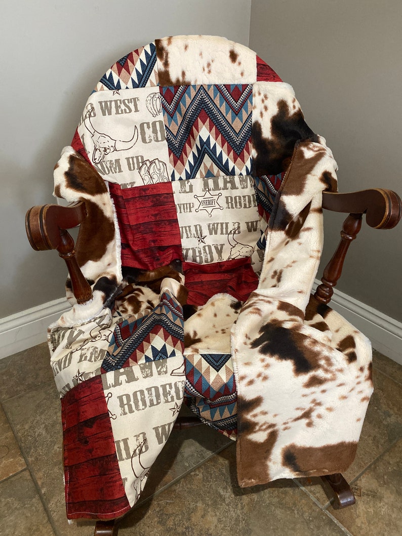 May include: A patchwork quilt with a cowboy theme. The quilt features a red, white, and blue color scheme with a variety of patterns, including cowhide, chevron, and woodgrain. The quilt is draped over a rocking chair.