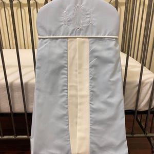 May include: A light blue baby crib bumper with a white center stripe and embroidered initials. The bumper is hanging on a metal crib frame. The initials are embroidered in white.
