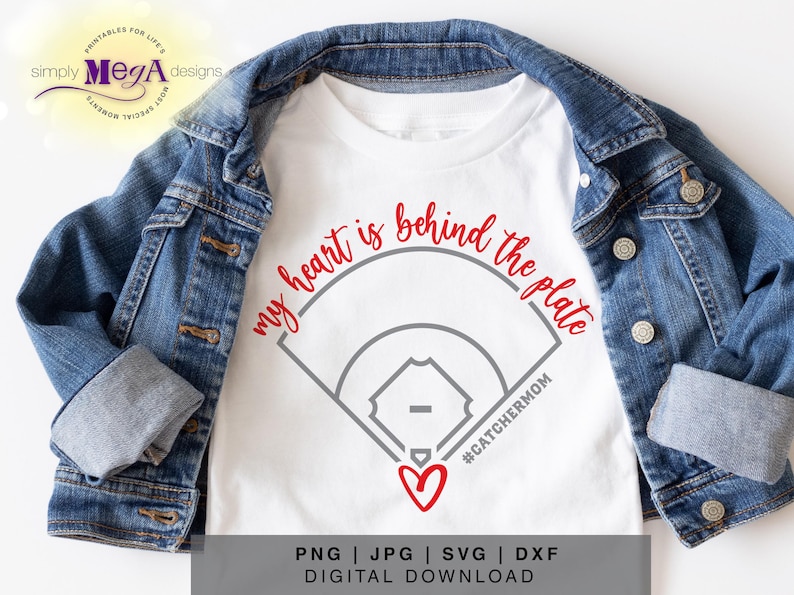 May include: White t-shirt featuring a baseball field graphic and the red text "my heart is behind the plate". The shirt also has the hashtag #CATCHERMOM and a red heart. A blue denim jacket is draped over the shirt.