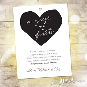 May include: A white card with a black heart shape and the text "a year of firsts". The card has a message about a basket of wine for a special pair, wishing them happiness and congratulating Alexa & Dominic. The card is signed "Love, Melissa & Liz" and has the text "simply Mega designs" at the bottom.