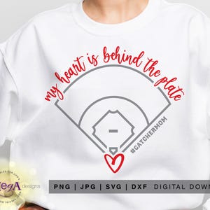 May include: White sweatshirt featuring the text "my heart is behind the plate" in red script above a grey baseball field graphic. A red heart and the hashtag #CATCHERMOM are below the graphic.