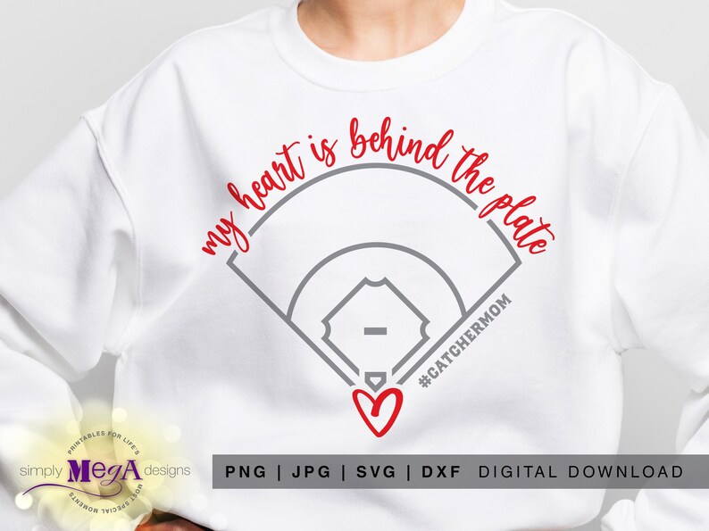 May include: White sweatshirt featuring the text "my heart is behind the plate" in red script above a grey baseball field graphic. A red heart and the hashtag #CATCHERMOM are below the graphic.