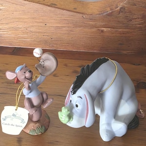 Peut inclure: Une figurine en céramique représentant un kangourou brun portant un t-shirt bleu et une casquette rouge, tenant un gant de baseball avec une balle sur un fil, à côté d'un âne gris regardant une petite grenouille verte. Une étiquette indique Catch the Spirit.