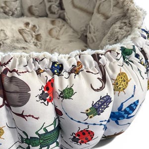 May include: A round pet bed featuring a white exterior fabric printed with a colorful pattern of various beetles and ladybugs. The interior is lined with a soft, cream-colored plush material that has a subtle embossed paw print texture.