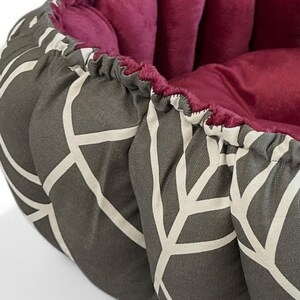 May include: A round pet bed with a grey geometric pattern on the exterior and a deep burgundy interior. The bed has a soft, cushioned design, suitable for small dogs or cats. Measurements are not visible.