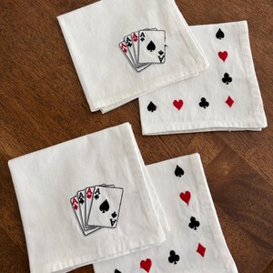 May include: Four white square fabric napkins featuring embroidered playing card designs. Two napkins display a fan of four aces, while the other two napkins feature a border pattern of red hearts, red diamonds, black spades, and black clubs.