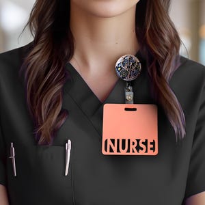 May include: A black scrub top with a peach-colored badge holder that reads "NURSE" in a cut-out design. A decorative badge reel with a steampunk design is attached. Two pens are in the pocket.