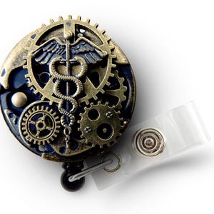 Puede incluir: Un portatarjetas de estilo steampunk con un s&iacute;mbolo m&eacute;dico de caduceo. El portatarjetas presenta una combinaci&oacute;n de colores azul y bronce con intrincados detalles de engranajes y una correa de pl&aacute;stico transparente.