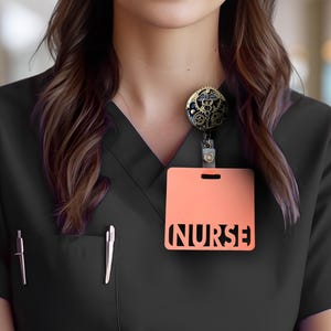 Puede incluir: Top de uniforme negro con un portatarjetas de color melocot&oacute;n. El portatarjetas tiene la palabra "NURSE" recortada. Se adjunta un carrete de insignia decorativo con engranajes y s&iacute;mbolos m&eacute;dicos. Dos bol&iacute;grafos est&aacute;n en el bolsillo.