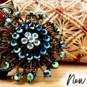 May include: A gold-toned brooch with a blue and iridescent crystal design. The brooch is shaped like a flower with multiple points. The word "New!" is written in black on a white background in the lower right corner of the image.