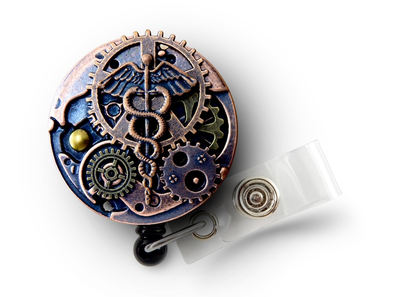 May include: A steampunk-style badge reel with a medical caduceus symbol. The round reel features bronze, copper, and blue gears and accents. A clear plastic strap and metal snap are attached for securing the badge.