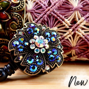 May include: A black retractable badge holder with a gold-toned, jeweled, floral design. The design features blue, purple, and clear rhinestones.